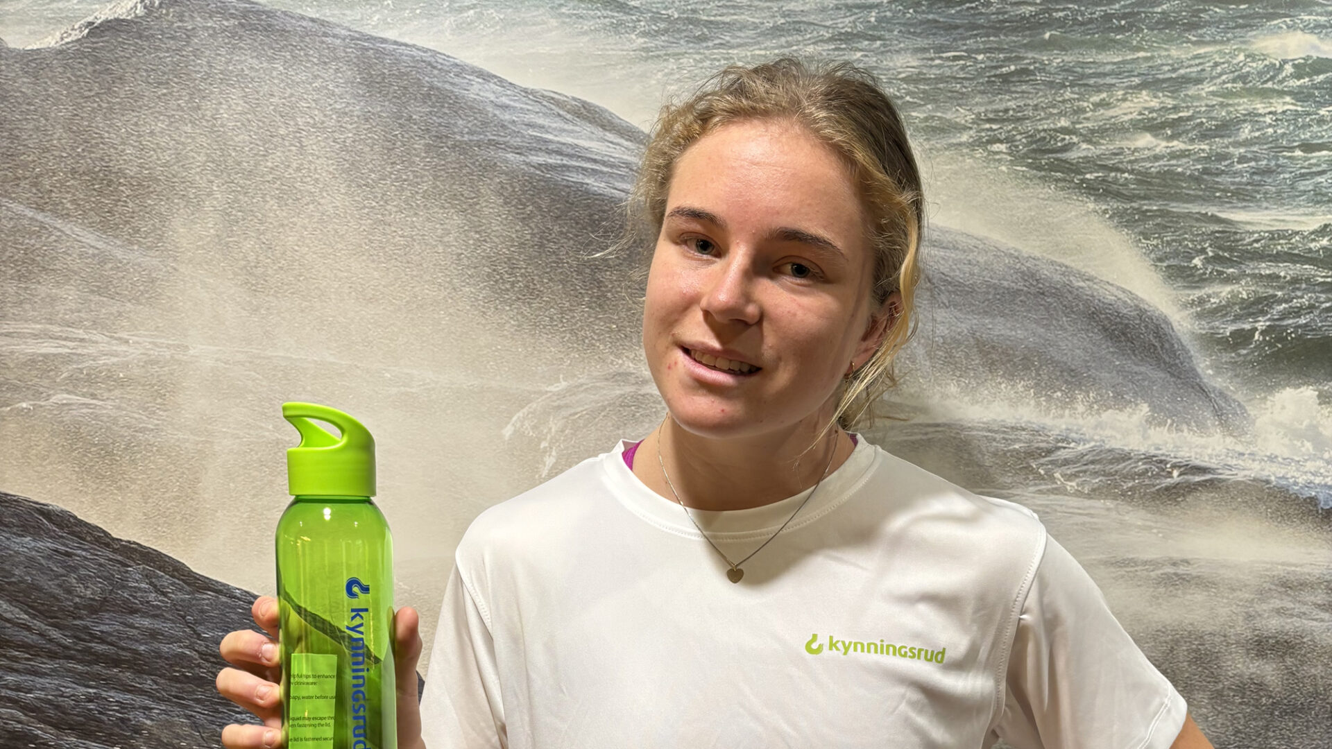 Ung kvinne i hvit t-shirt som er dyktig i friidrett poserer foran et bilde av havet og med en grønn drikkeflaske i hånda.